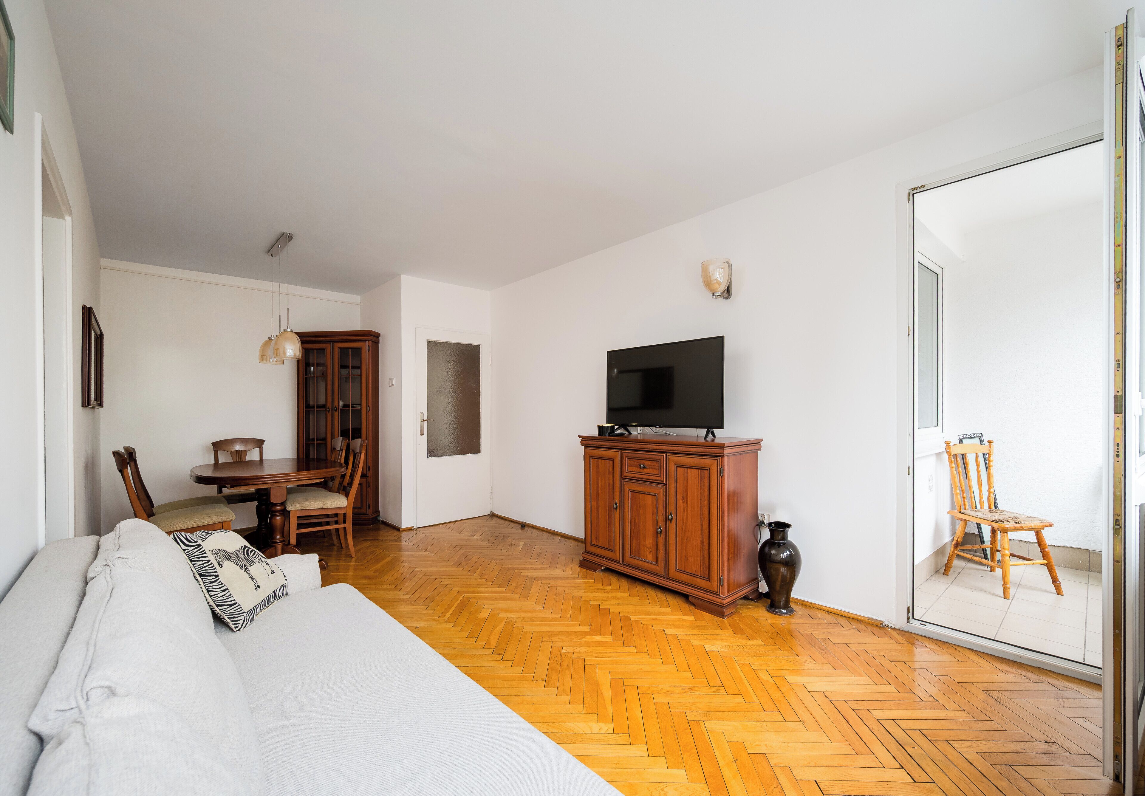 Une pièce spacieuse dotée d’un parquet en bois disposé en motif à chevrons, d’un grand lit, d’un téléviseur posé sur un meuble en bois, d’une table à manger avec des chaises et d’une grande fenêtre.