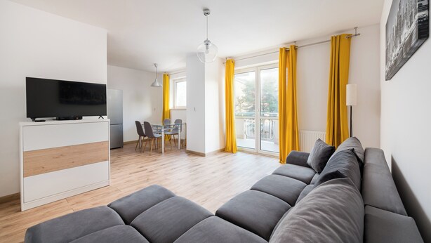 A modern living room with a grey sofa, a flat-screen TV on a white cabinet, a dining area with chairs, and a balcony with yellow curtains. - Bemowo Lotnisko Snug Apartment (Warszawa)