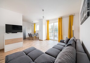 A modern living room with a grey sofa, a flat-screen TV on a white cabinet, a dining area with chairs, and a balcony with yellow curtains. - Bemowo Lotnisko Snug Apartment (Warszawa)