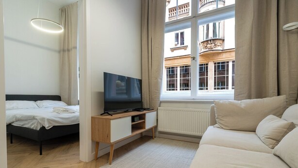 A modern bedroom with a bed, a TV, a sofa, and a window with a view of buildings. - Dyrekcyjna Charming Apartment (Katowice)
