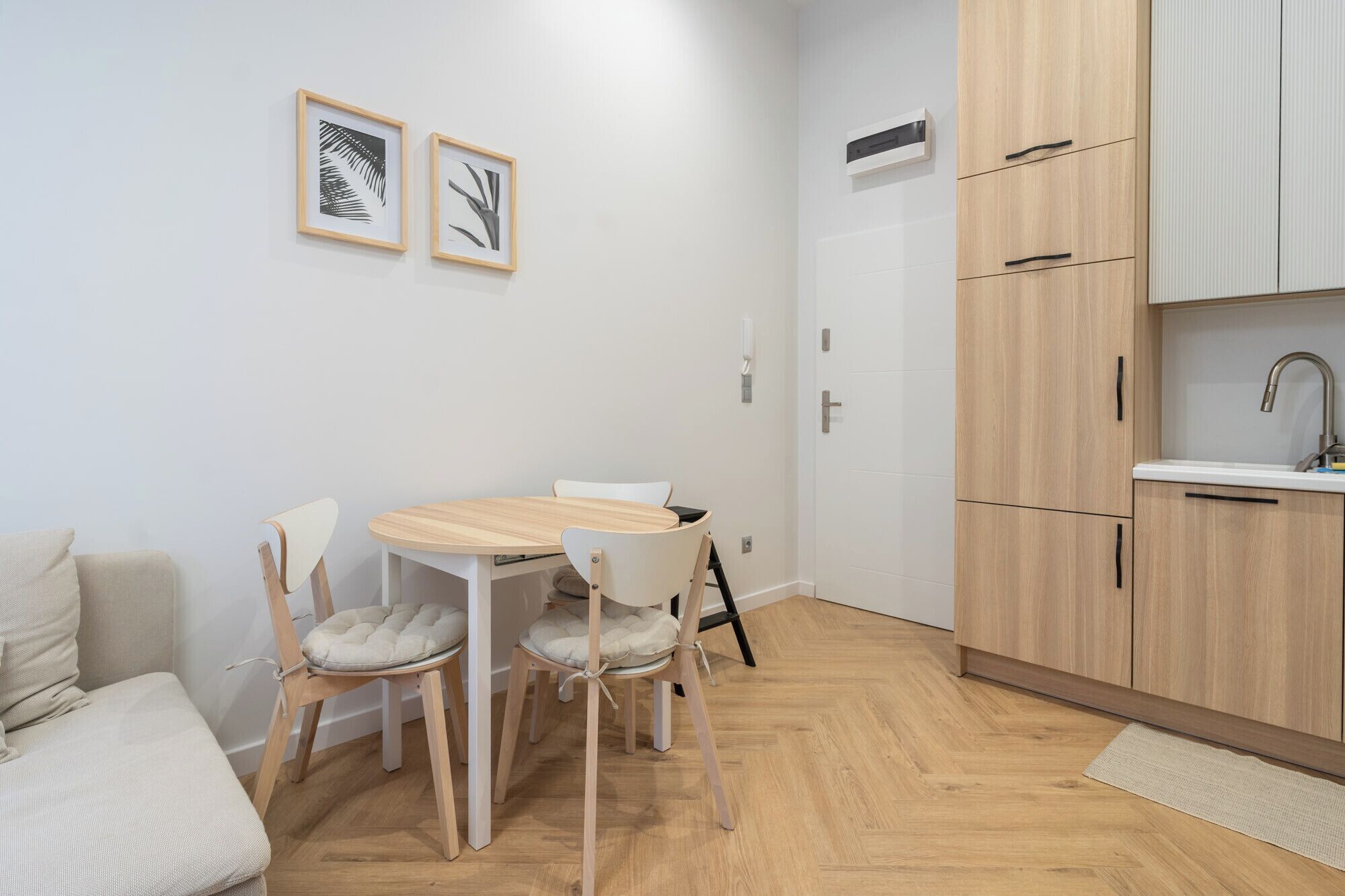 Et kompakt køkken med træskabe, en hvid bordplade og en vask. Der er et spisebord med stole og en lille sofa i opholdsstuen.