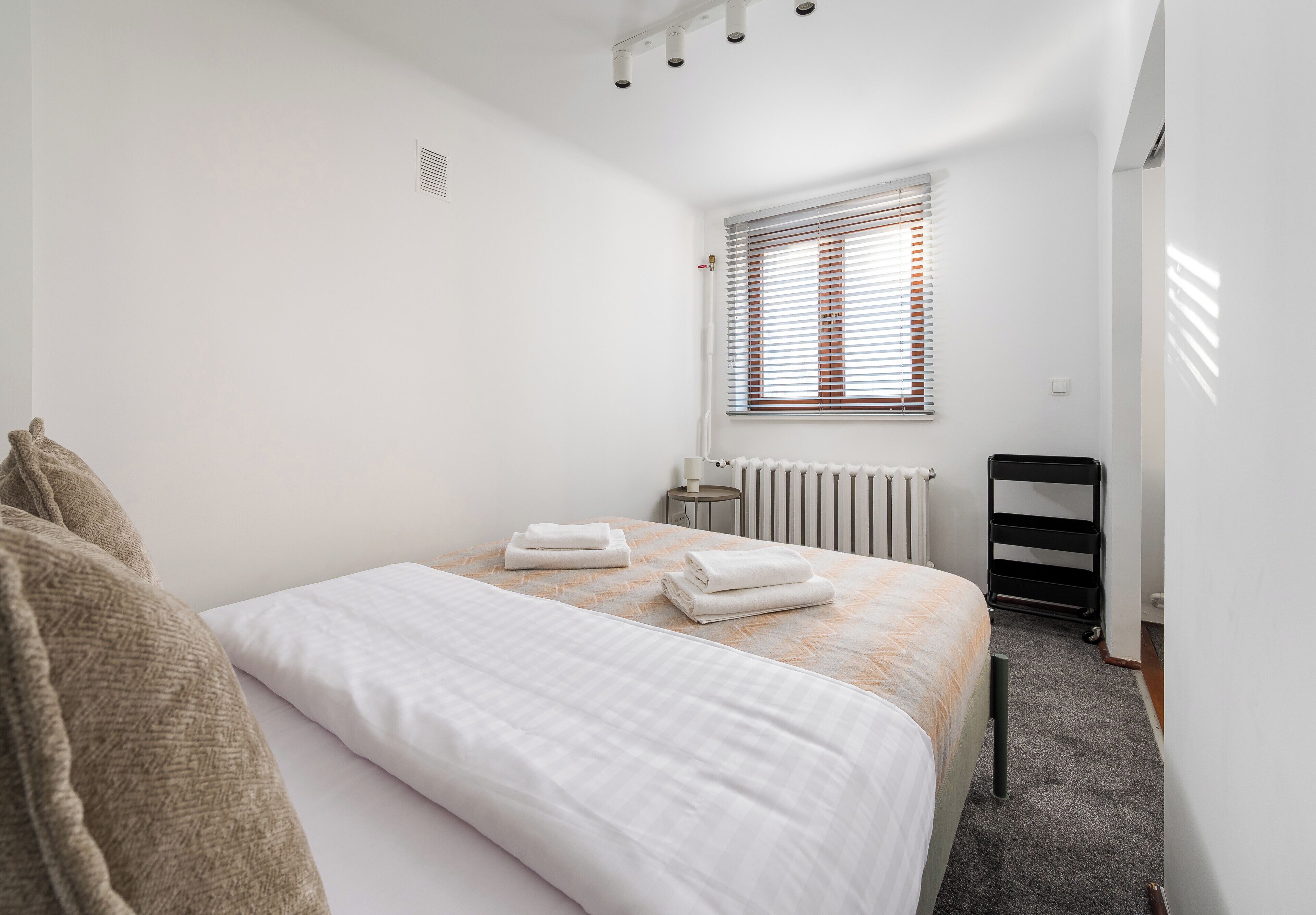 A bedroom with a bed, a radiator, a window with blinds, and a bedside table.