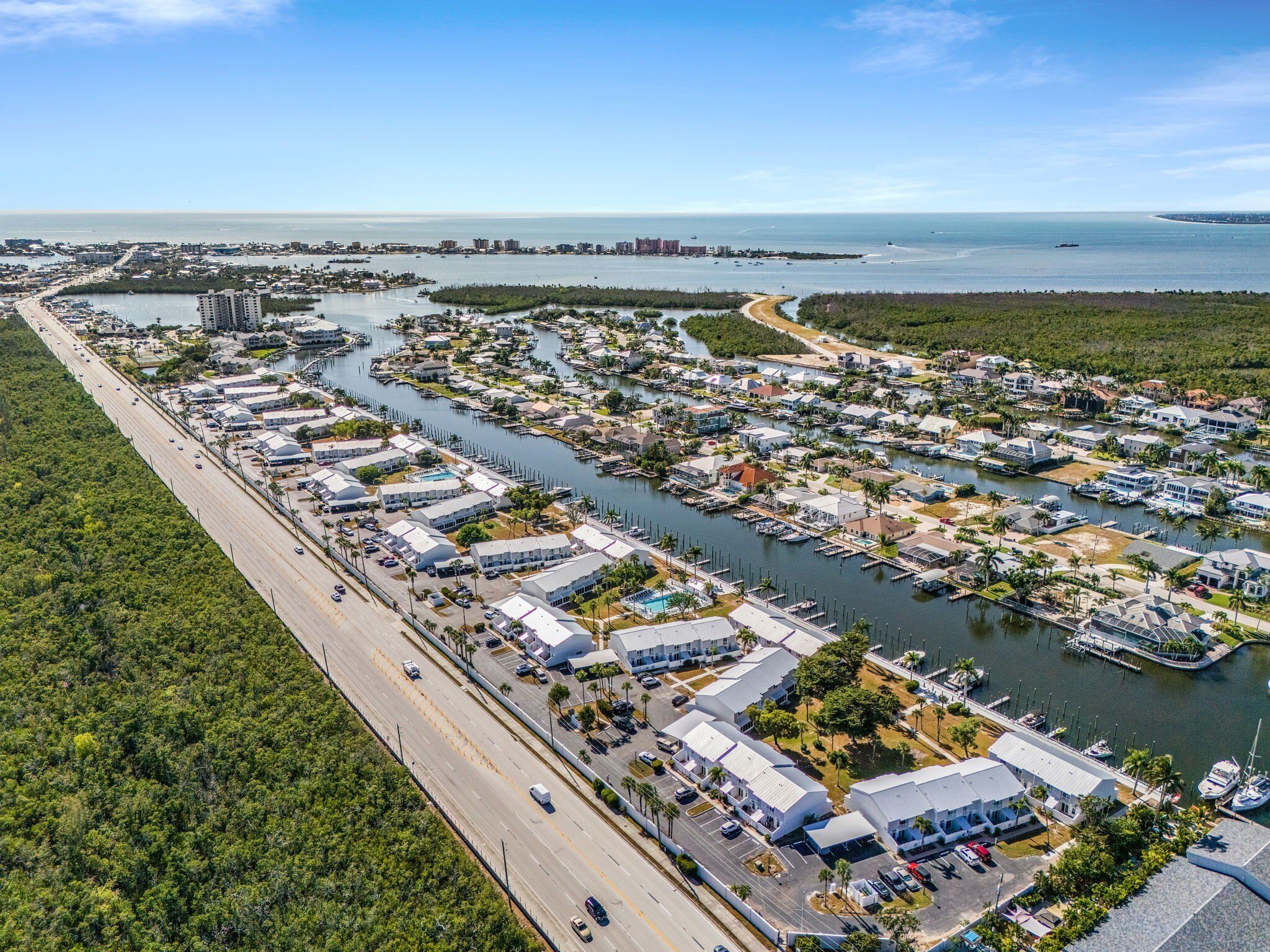 Vue aérienne d’un quartier résidentiel situé sur la côte, avec un port de plaisance, une route parallèle à la mer et des bâtiments dotés de toits plats.