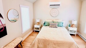Une chambre avec un grand lit, deux tables de chevet, un banc et un appareil de climatisation fixé au mur.