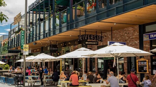 Un espace extérieur aménagé pour les repas, avec des tables et des parasols, un bâtiment en briques sur lequel on peut lire l’inscription « RIVERSIDE MARKET », et un bâtiment moderne portant l’enseigne « KAISER BREWERY ».