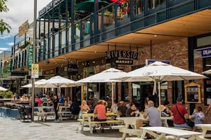 An outdoor dining area with tables and umbrellas, a brick building with a sign reading "RIVERSIDE MARKET", and a modern building with a sign for "KAISER BREWERY". - Modern CBD Studio w/ King Bed | Ground Floor Ease (Christchurch)