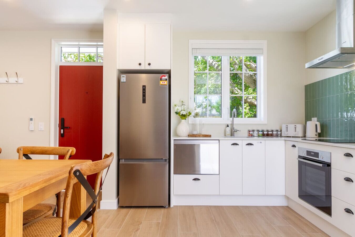 A modern kitchen with white cabinets, stainless steel appliances, a red door, and a window with a view of greenery.