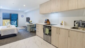 A modern kitchen with light wood cabinets, a built-in oven, and a sink. There's a dining area with a table and chairs, and a bed with a white duvet. - Modern Studio Near Stadium, Dining & City Fun (Christchurch)