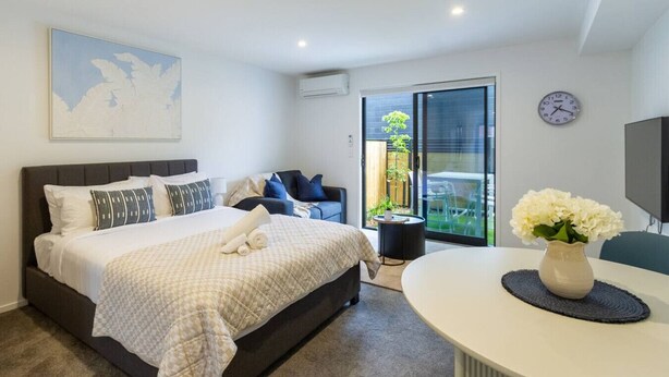 A modern bedroom with a large bed, a bedside table, a chair, and a view of the outdoors through a sliding glass door. - Modern Studio Near Stadium, Dining & City Fun (Christchurch)