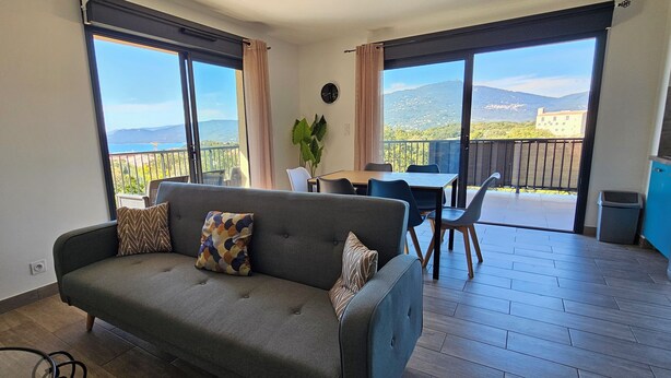 A modern living room with a sofa, dining table, and chairs, a balcony with a view of the sea and mountains, and a kitchen area. - T3 Le Myrte sea & mountain view ideally located (PROPRIANO)
