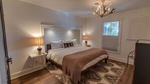 A bedroom with a large bed, bedside lamps, a chandelier, and a window with blinds. - Lofts on Main - Ravenel (Highlands)