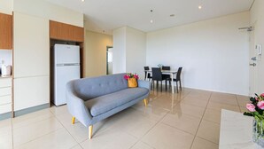Ein modernes Wohnzimmer mit grauem Sofa, einem Esstisch mit Stühlen, einer Küchenzeile inklusive Kühlschrank und einer Tür zu einem weiteren Raum.