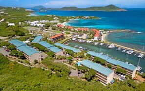 An aerial view of a coastal resort with multiple buildings, a marina with boats, and a clear view of the surrounding ocean and hills.