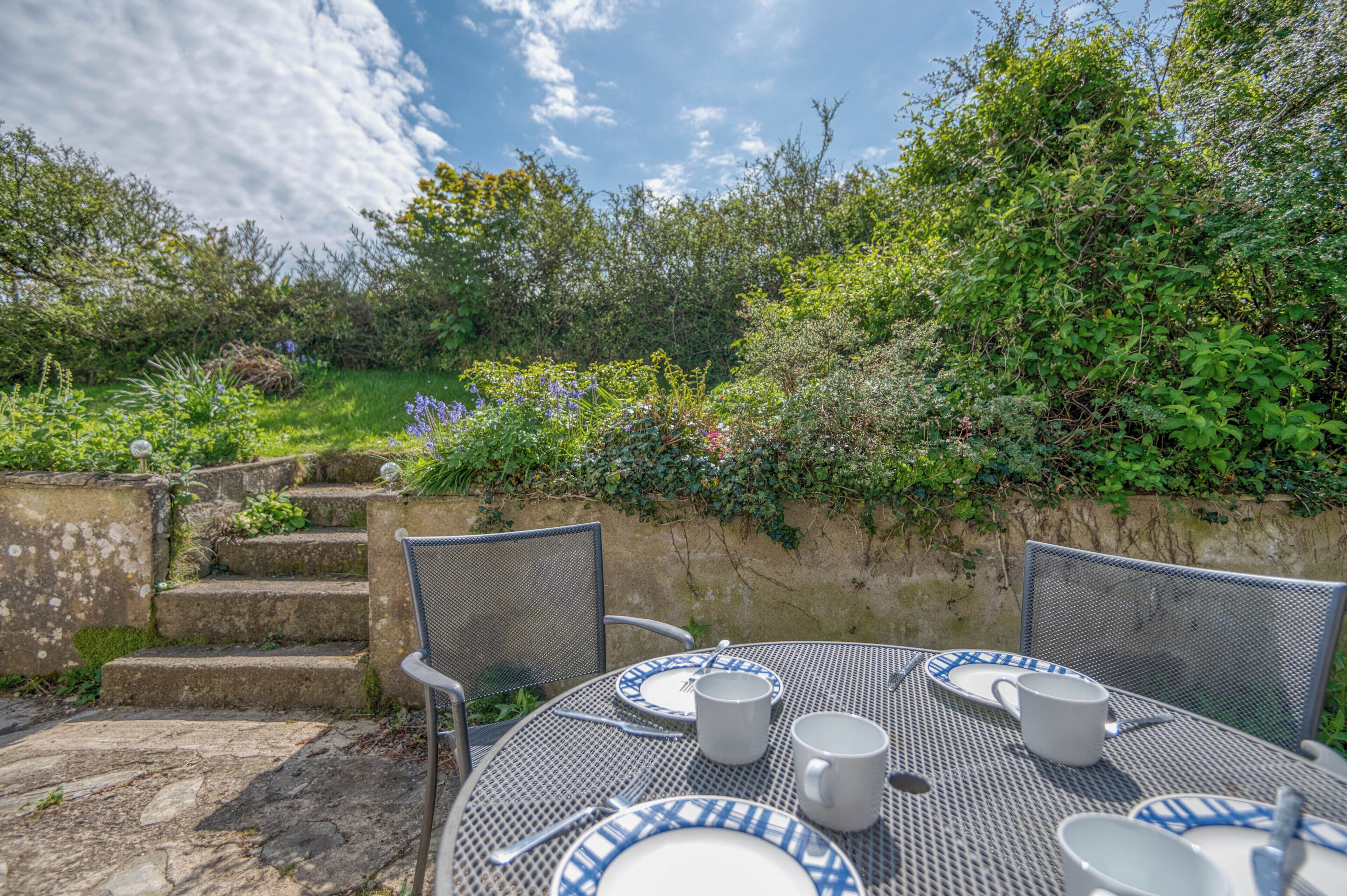 Une terrasse avec une table dressée pour deux personnes, entourée de verdure et bordée d’un mur de pierres.