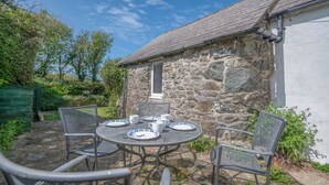 Un bâtiment en pierre avec une petite fenêtre, une table ronde en métal entourée de chaises, et une clôture verte en arrière-plan.