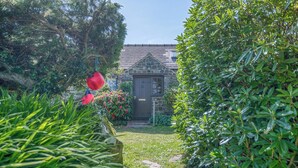 Un bâtiment en pierre avec une porte sombre, entouré d’une végétation luxuriante et bordé d’un chemin en pierre.