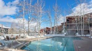 A pool area with a clear blue pool, surrounded by snow-covered trees and buildings. - Enclave 313 | Ski-In/Ski-Out Condo w/Shared Pool/Hot Tub/Gym/WiFi (Snowmass Village)