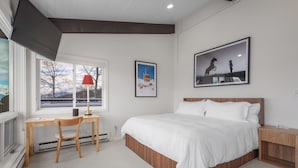 A bedroom with a large bed, a desk with a chair, a window with a view of trees, and a framed picture on the wall. - Enclave 313 | Ski-In/Ski-Out Condo w/Shared Pool/Hot Tub/Gym/WiFi (Snowmass Village)