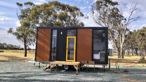 Una cabaña pequeña y moderna con fachada de madera y puerta amarilla, que cuenta con un pequeño patio con mesa y sillas.