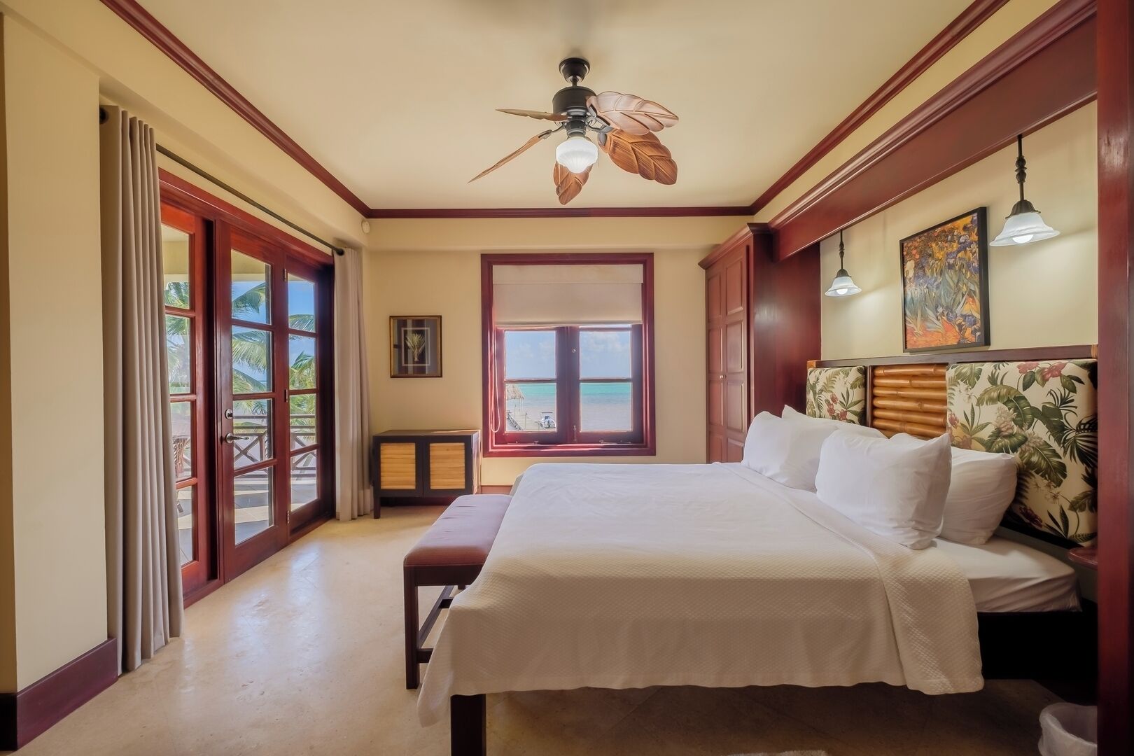 A hotel room with a bed, a ceiling fan, a window with a view of the beach, and a door leading to a balcony.
