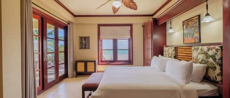 A hotel room with a bed, a ceiling fan, a window with a view of the beach, and a door leading to a balcony.