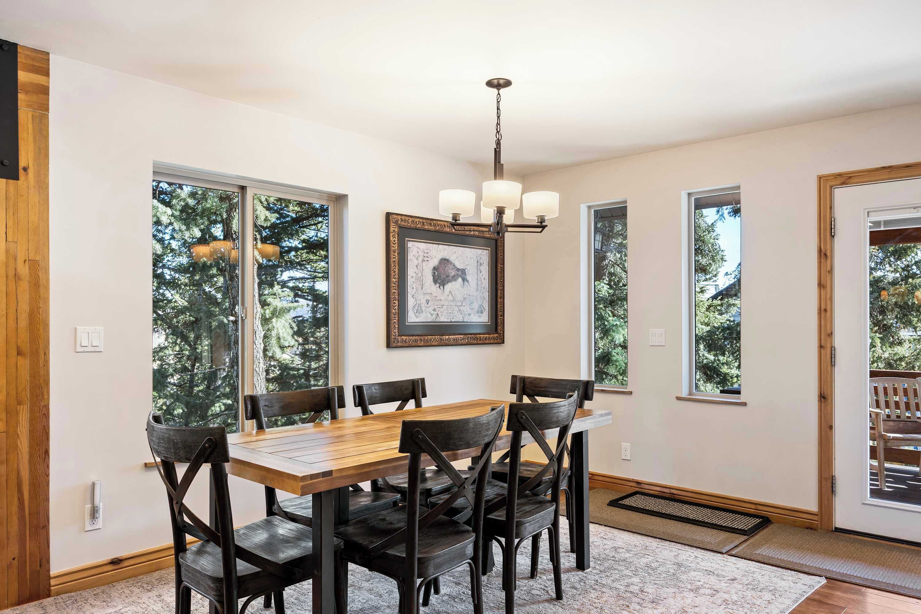 Un coin repas avec une table et des chaises en bois, de grandes fenêtres donnant sur la verdure, un lustre et un tableau encadré au mur.