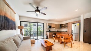 A modern living room with a sofa, coffee table, and dining table with chairs. There is a kitchen area with a refrigerator, microwave, and cabinets. A ceiling fan is visible, and the room has large windows. - Depa Moderno con Alberca Tulum 1 Rec (Tulum)