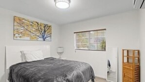 Une chambre à coucher avec un lit, une fenêtre avec des stores, un tableau au mur et une armoire en bois.
