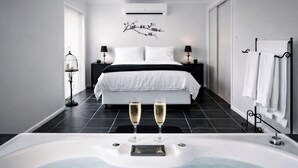 Une chambre moderne avec un grand lit, une table de chevet avec une lampe, un climatiseur fixé au mur et une baignoire avec deux coupes de champagne.
