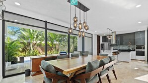 Eine moderne Küche mit einer großen Kochinsel, einem Essbereich mit Holztisch und Stühlen sowie großen Fenstern mit Blick auf Grünfläche.