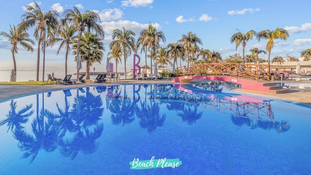 Ein Pool mit Palmen und Liegestühlen, eine Brücke und ein Schild mit der Aufschrift 'Beach Please'.