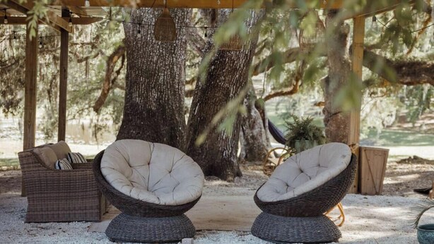 Dos sillas de mimbre con asientos acolchados bajo una pérgola de madera, rodeadas de árboles.