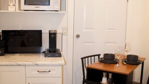 A compact kitchen with white cabinetry, a microwave, a coffee maker, and a dining area with a wooden table and chairs. - Studio - Cozy Downtown Hideaway (Greater Sudbury)
