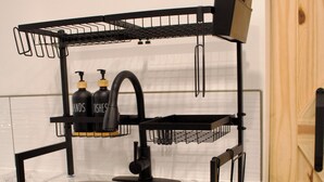 A kitchen with a double sink, black faucet, and a black wire rack above the sink. There are two bottles of hand soap on the rack. - Studio - Cozy Downtown Hideaway (Greater Sudbury)