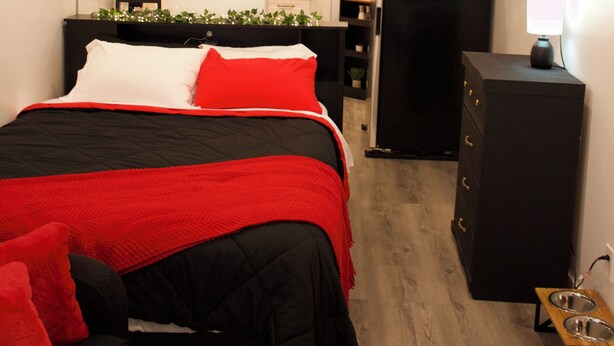A bedroom with a bed, a TV mounted on the wall, a clock, and a small kitchenette area. - Studio - Cozy Downtown Hideaway (Greater Sudbury)