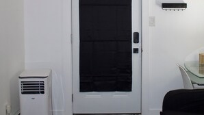 A room with a white door, a black mat at the entrance, a white air conditioner, and a black sofa. - Studio - Cozy Downtown Hideaway (Greater Sudbury)