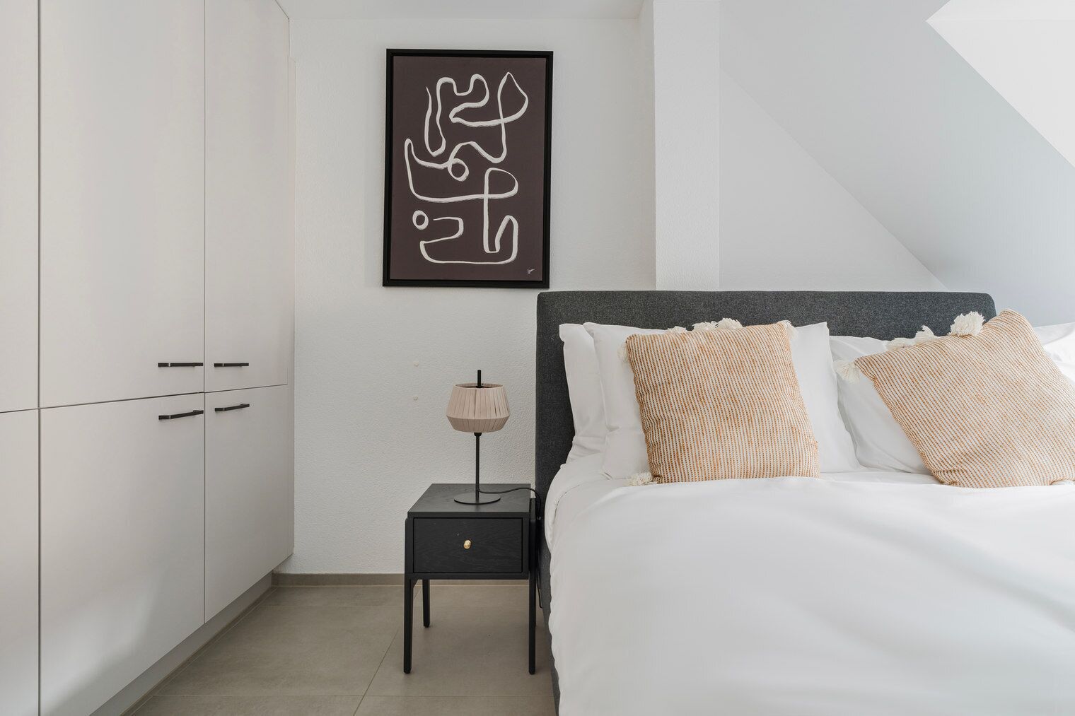 Een slaapkamer met een bed, nachtkastje, lamp en een lijst met abstracte kunst aan de muur.