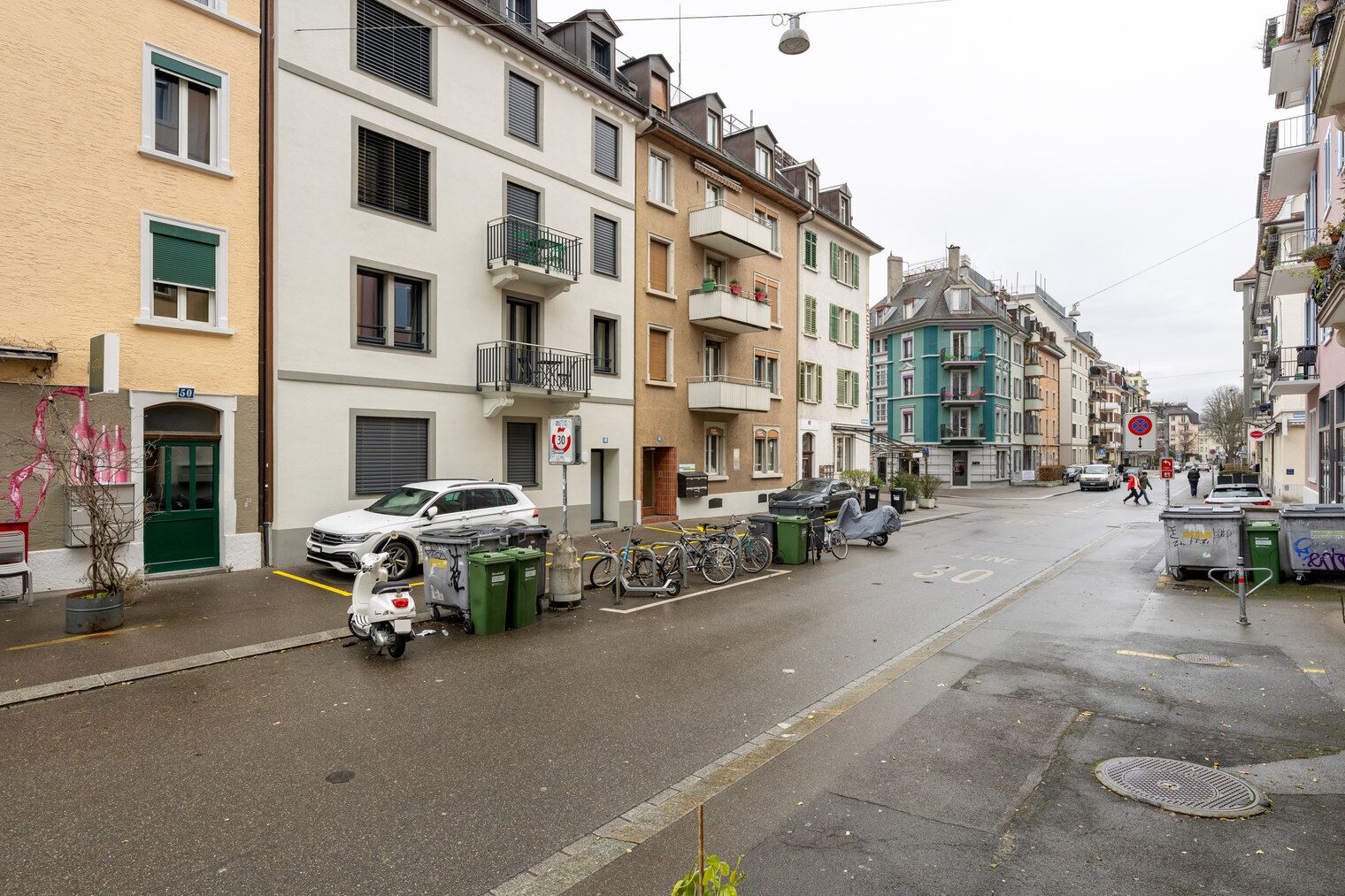 Een straat met geparkeerde auto's en fietsen, gebouwen met balkons, en een vrijliggende weg.