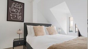 A modern bedroom with a sloped ceiling, a bed with white bedding and two beige pillows, a small bedside table with a lamp, and a framed abstract artwork on the wall. - Blueground | Sihlfeld, washer/dryer, nr Zürich HB (Zurich)