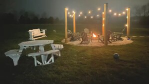 A covered outdoor seating area with a fire pit, string lights, and picnic tables.