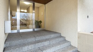 Kivestä rakennettu porrassarja johtaa lasiovelle, ja sen takaa avautuu näkymä rakennukseen ja sen sisällä olevaan kasviin.