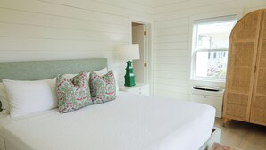 Een keurig opgemaakt bed met witte lakens en sierkussens met bloemenmotief, een nachtkastje met een groene lamp, en een rieten kast.