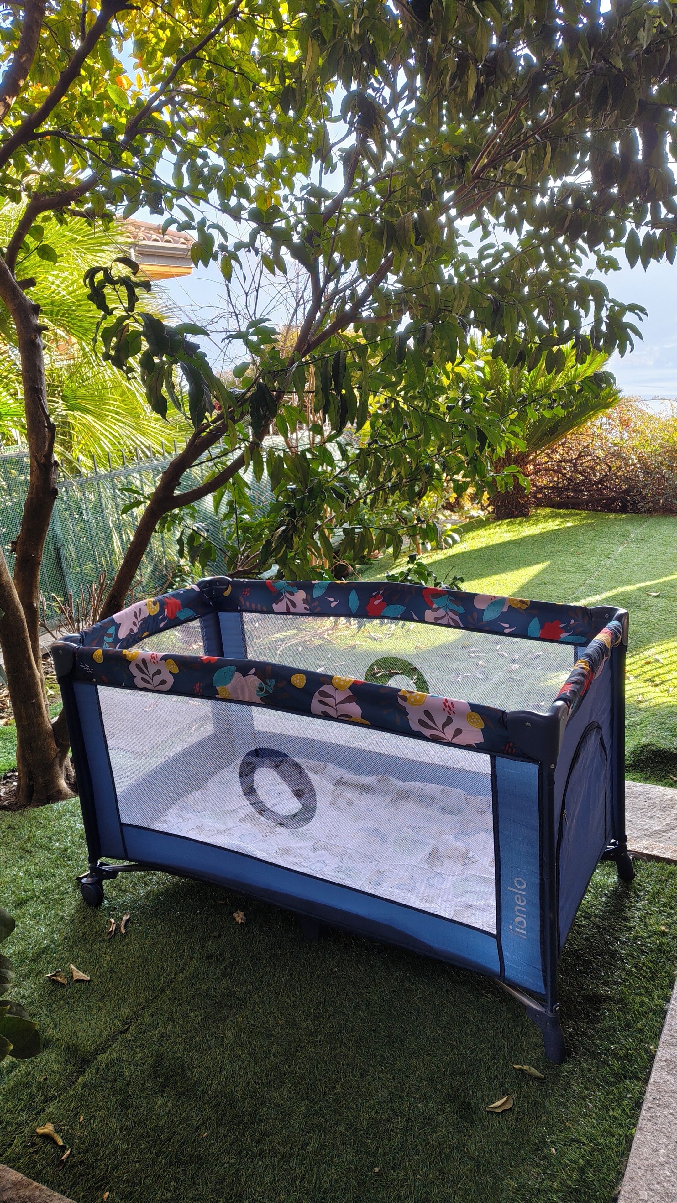 A playpen with a floral pattern, placed on artificial grass, surrounded by trees and a fence.
