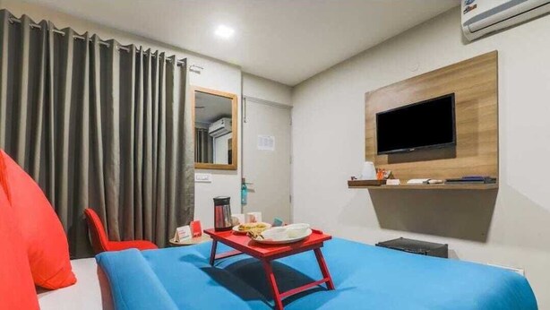 A hotel room with a bed, a red table with a plate and a cup, a TV mounted on the wall, and a window with curtains. - Loginn Stays #1 (Gurugram)