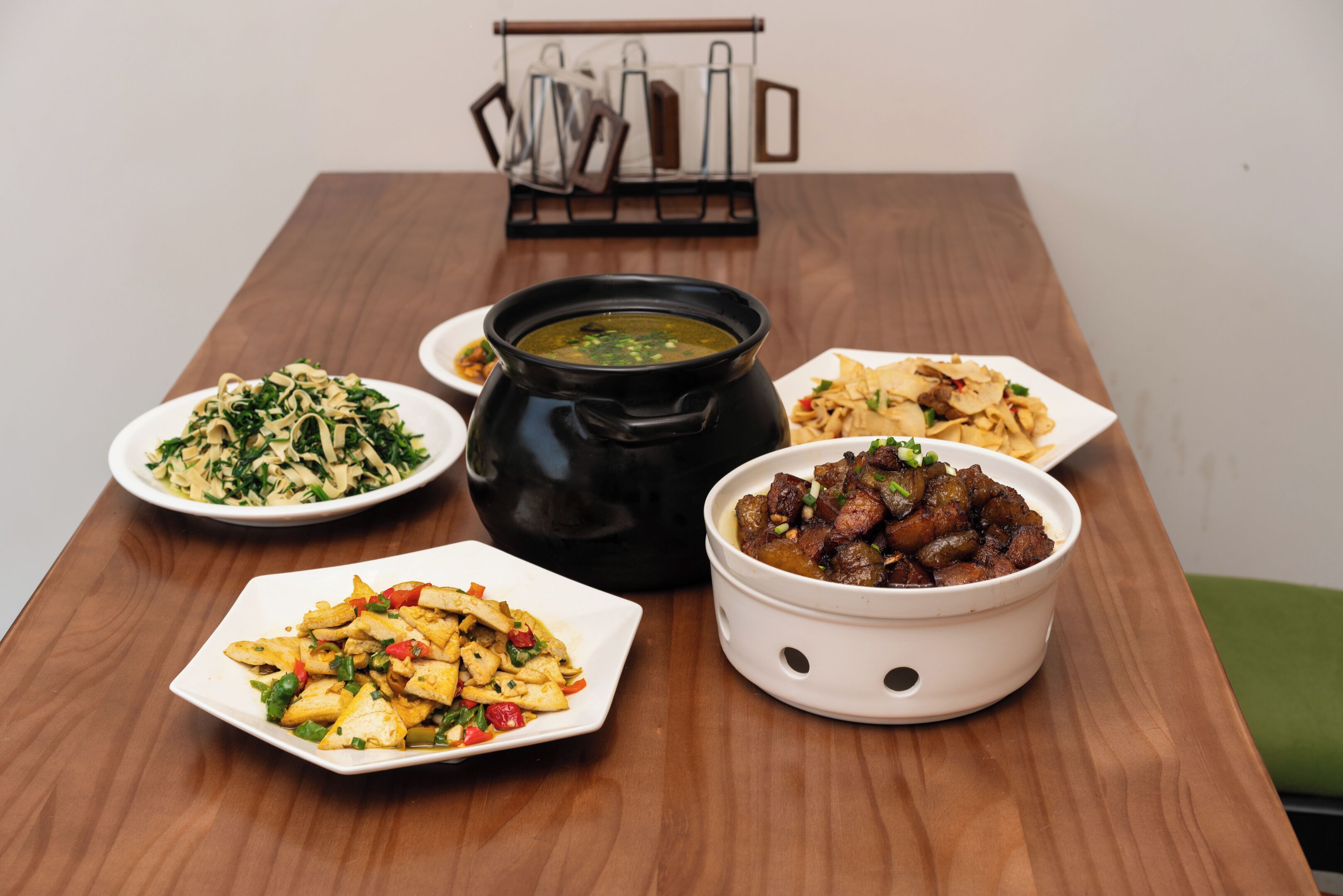 A wooden table with various dishes including a bowl of soup, a plate of noodles, a bowl of meat, and a plate of stir-fried vegetables.
