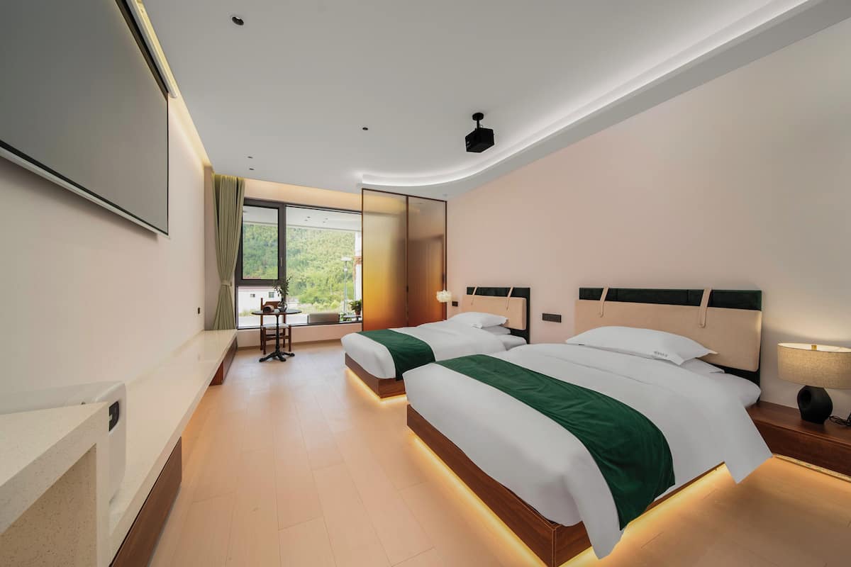 A modern hotel room with two beds, a large window, a desk, and a mirror.