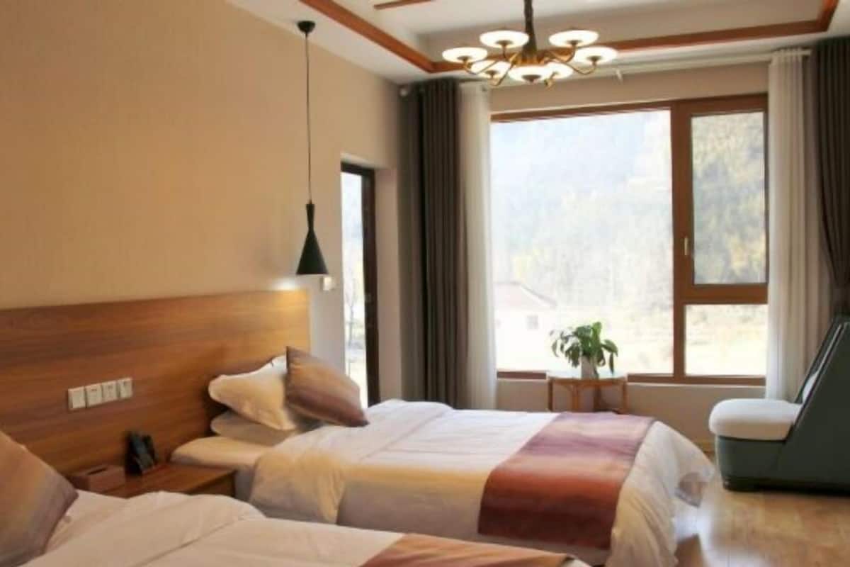 A hotel room with two beds, a wooden headboard, a small table with a plant, and a window with curtains.