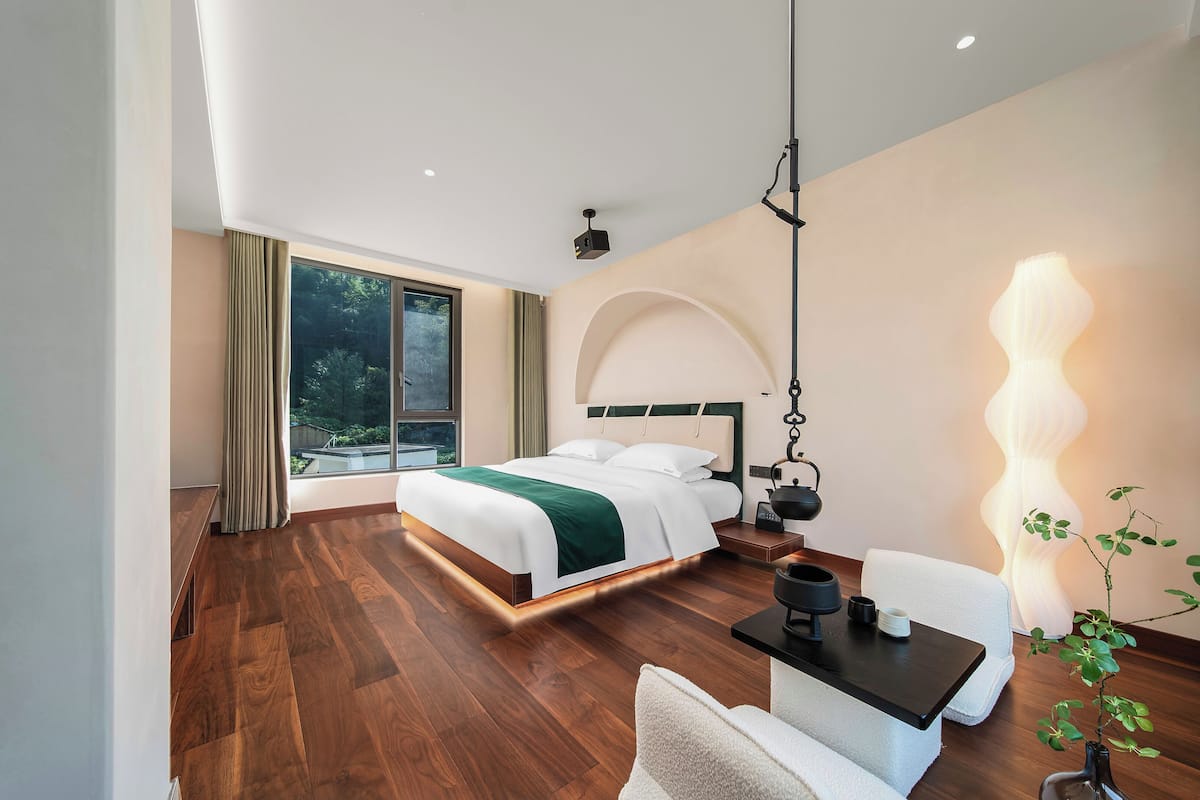 A modern hotel room with a large bed, a desk, and a view of greenery outside.