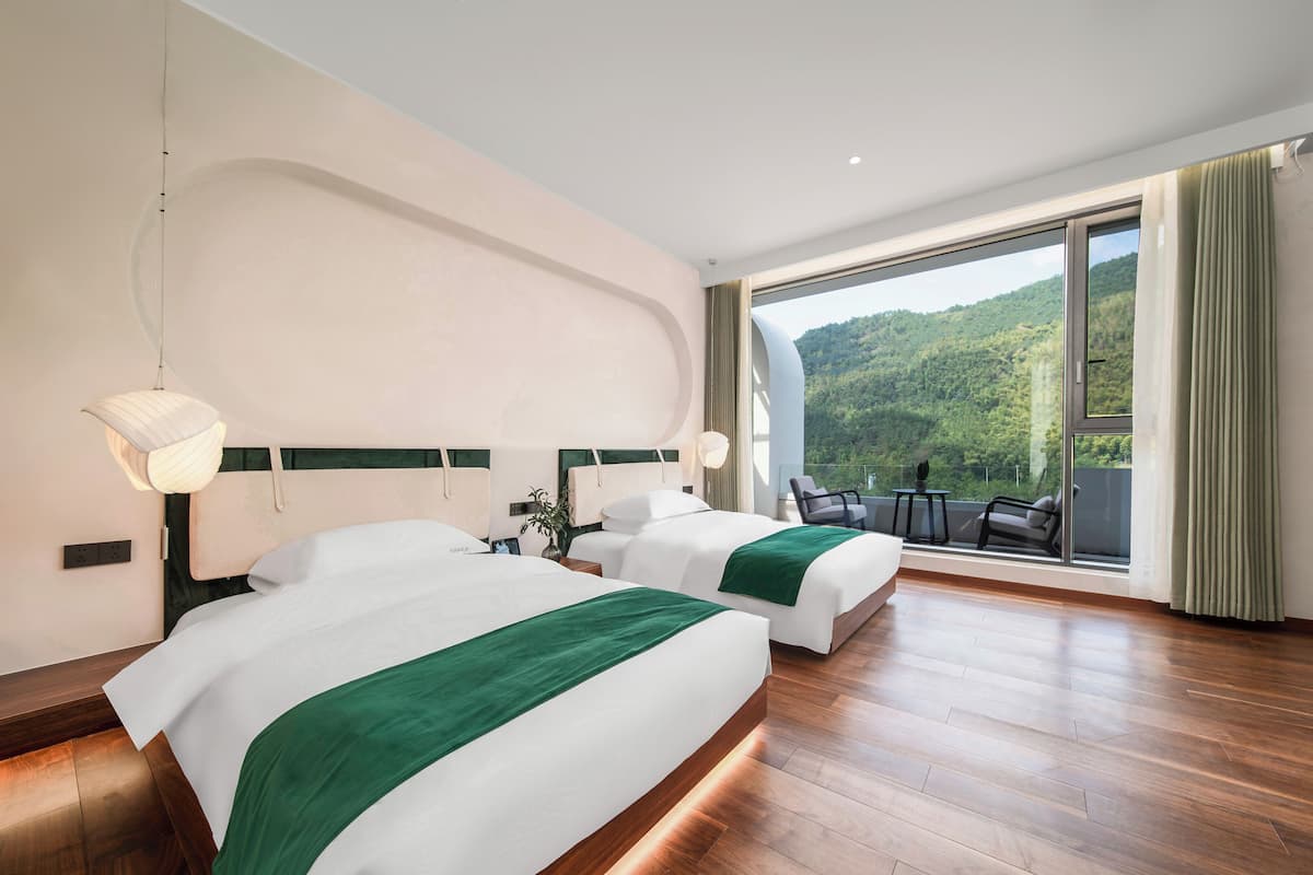 A hotel room with two beds, a balcony with a view of a forested hillside, and a modern design.