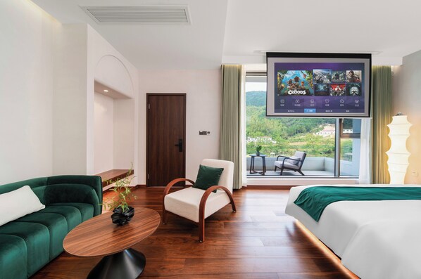 A hotel room with a large TV displaying a movie selection, a green sofa, a wooden coffee table, a white bed, and a view of a lush landscape through large windows. - Bright Moon Valley Smart Hotel (Anqing)